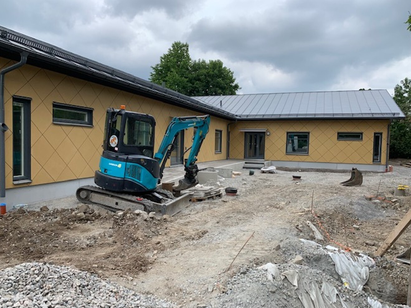 Grävmaskin i arbete framför en gul förskolebyggnad i ett plan med grått tak. 