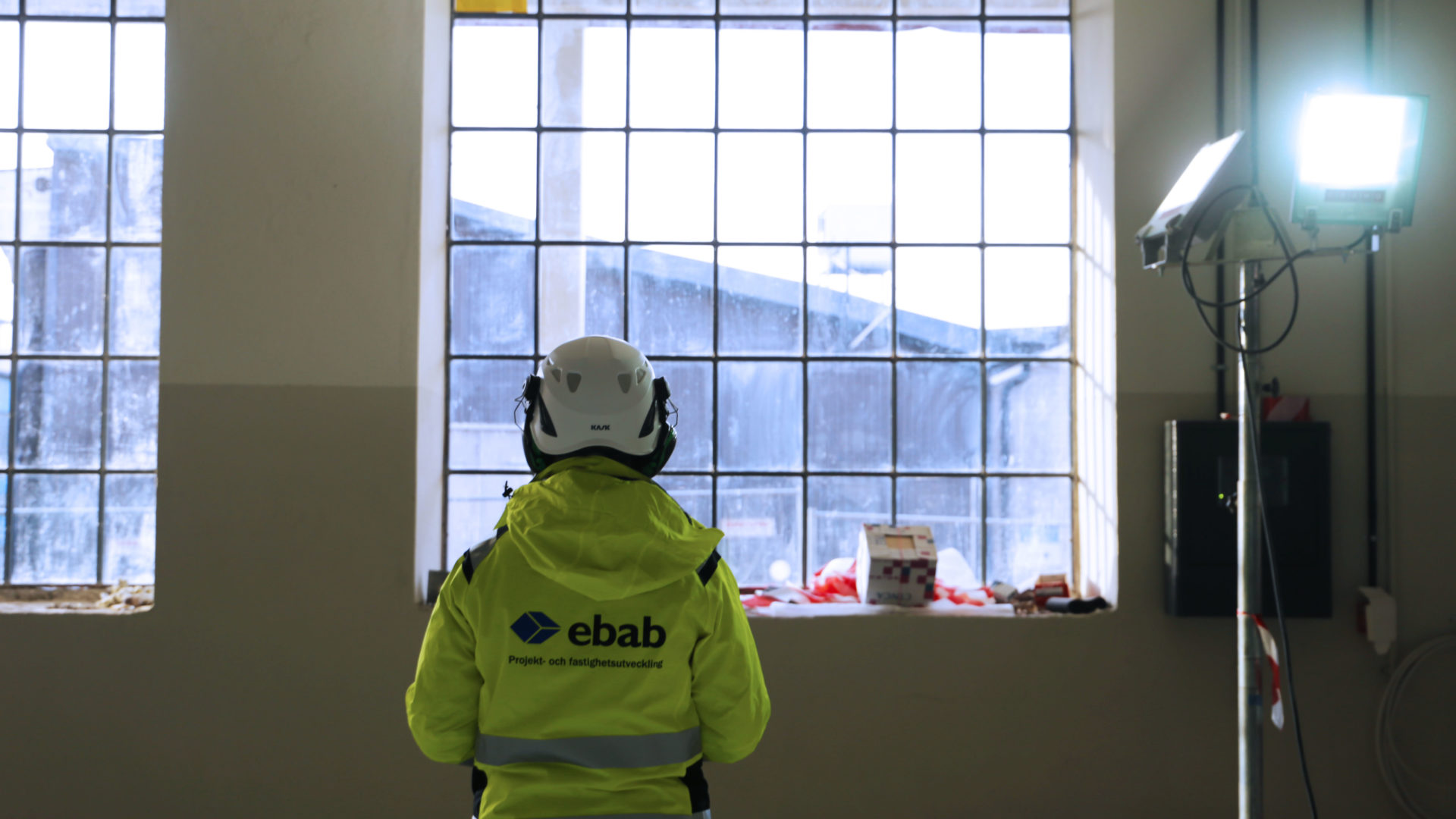 Ryggen på en person med hjälm och gul jacka med Ebab-tryck med ett fönster i bakgrunden. Hos Ebab får du möjlighet att utvecklas.