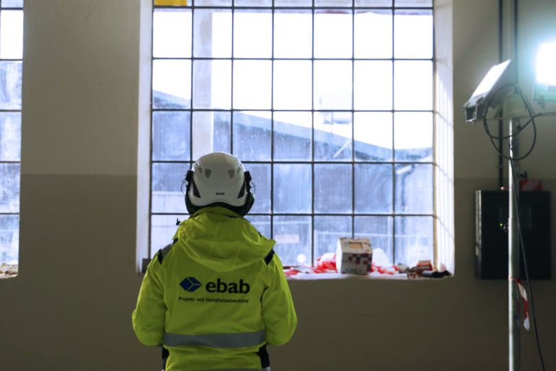 Ryggen på en person med hjälm och gul jacka med Ebab-tryck med ett fönster i bakgrunden. Hos Ebab får du möjlighet att utvecklas.