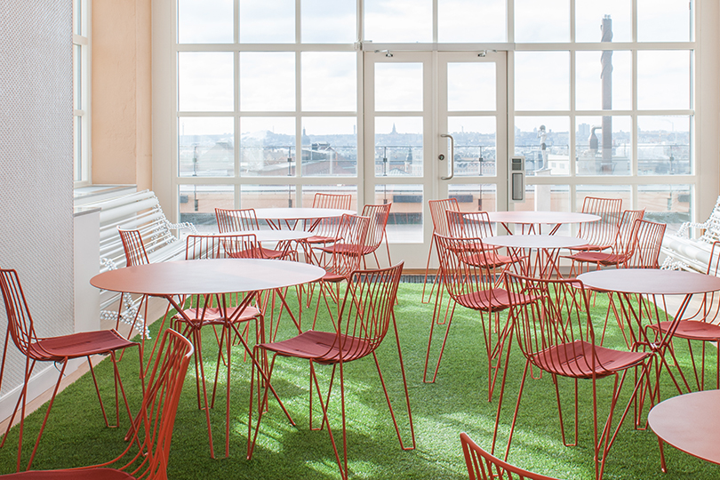 En matsal med grön heltäckningsmatta och röda metallmöbler mot ett stort fönster i bakgrunden.